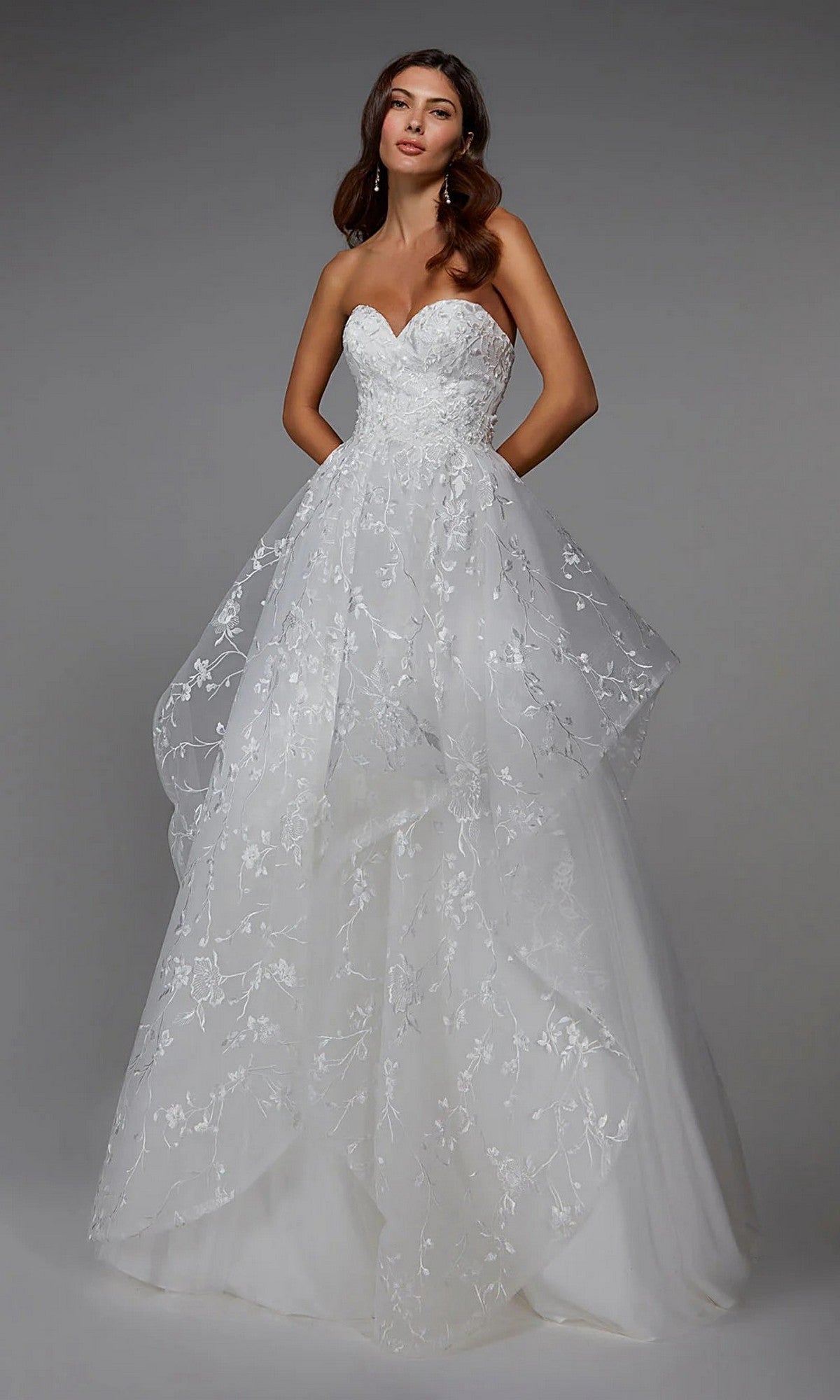 A woman models the Alyce 7041 long formal dress in ivory, featuring a strapless tulle ball gown design with floral lace details and a voluminous layered skirt, against a plain grey background.